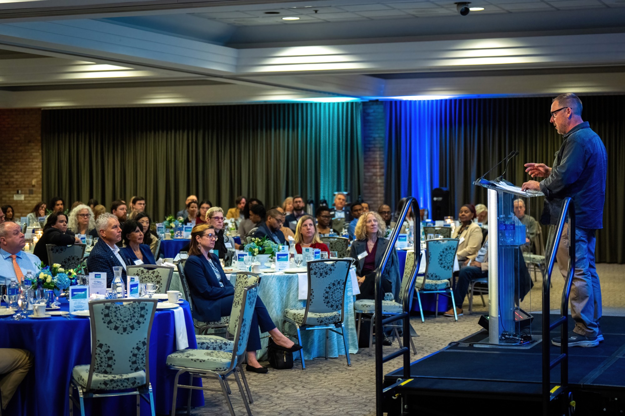 A speaker at the podium from the side with several dinner tables in view